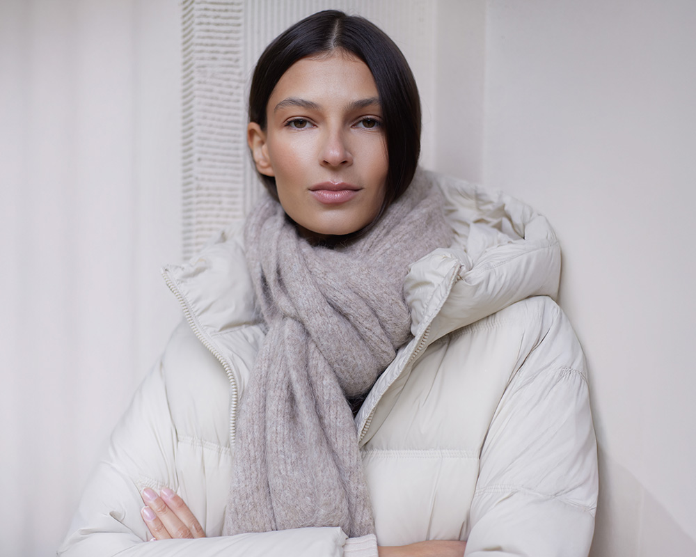 Kobieta w jasnej kurtce puchowej i szaliku stoi oparta o ścianę.