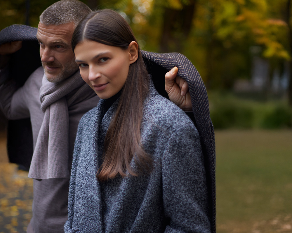 Kobieta i mężczyzna w eleganckich szarych płaszczach i szalikach stoją w parku.