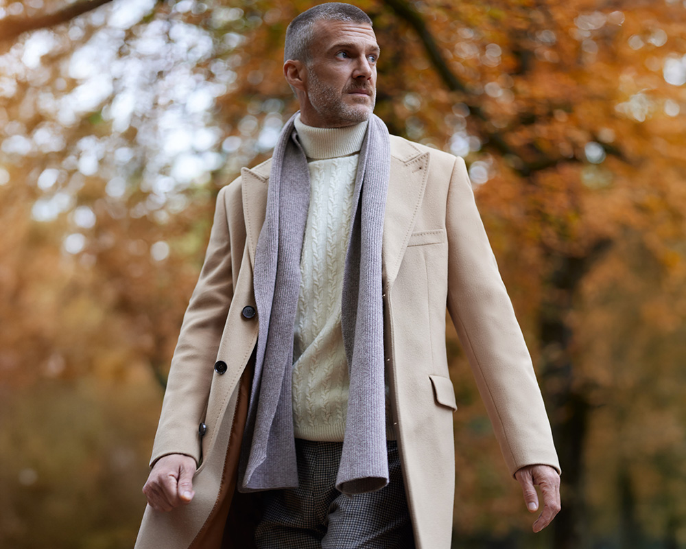 Mężczyzna w beżowym płaszczu i szaliku spaceruje w jesiennym parku.
